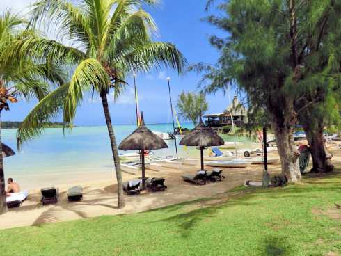 Tropischer Strand auf Mauritius mit Palmen, Sonnenliegen unter Strohdächern und Segelbooten am türkisfarbenen Wasser.