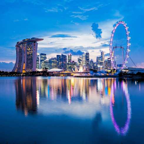 Beleuchtete Skyline von Singapur bei Abenddämmerung mit dem Singapore Flyer, Marina Bay Sands und farbenfroher Spiegelung im Wasser.