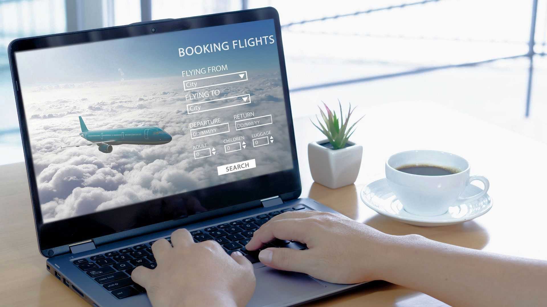 Person bucht Flug online auf einem Laptop mit Kaffee und Pflanze im Hintergrund.