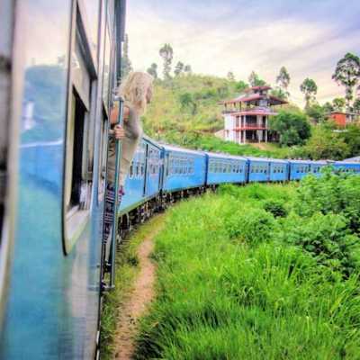 Frau lehnt aus einem blauen Zugfenster bei einer Zugfahrt durch das grüne Hochland von Sri Lanka