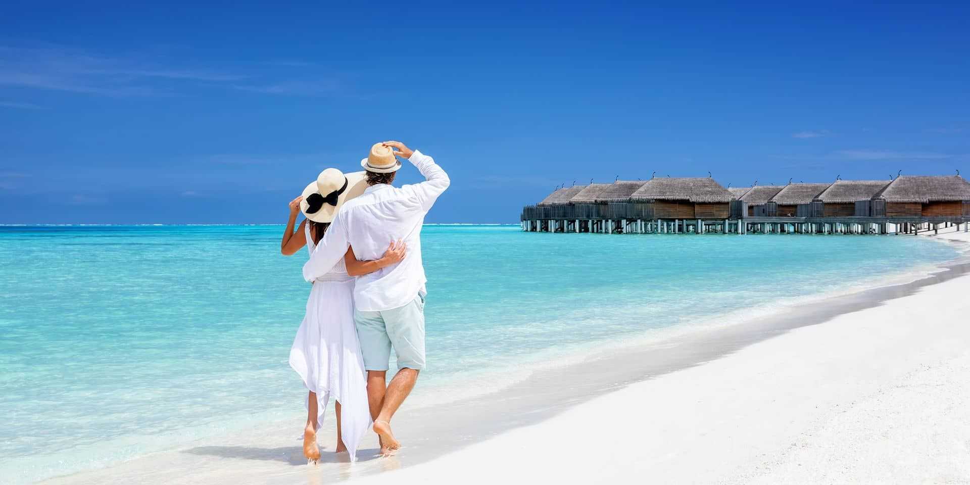 Paar in weißer Kleidung am Strand der Malediven, blickt auf Wasserbungalows
