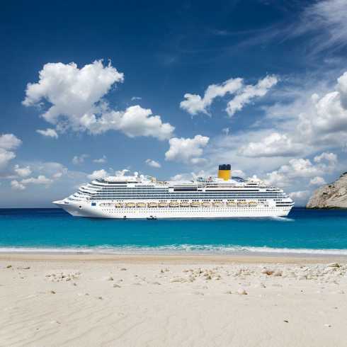 Großes Kreuzfahrtschiff vor weißem Sandstrand im türkisblauen Meer unter blauem Himmel mit Wolken.
